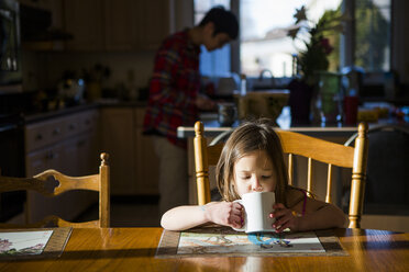 Tochter trinkt Tee auf dem Tisch, während der Vater im Hintergrund zu Hause arbeitet - CAVF53693