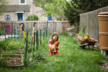 Mädchen spielt mit Huhn, während sie auf einer Wiese im Hof kniet - CAVF53694