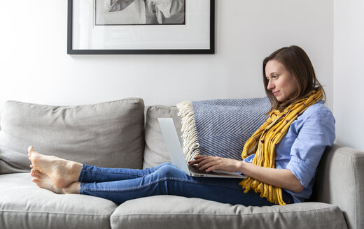 Seitenansicht einer Frau, die einen Laptop benutzt, während sie auf dem Sofa im Wohnzimmer sitzt - CAVF53733
