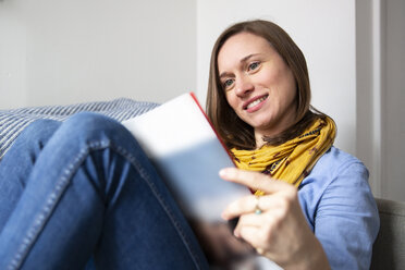 Smiling woman reading magazine while sitting on sofa at home - CAVF53734