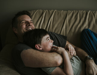 Happy father embracing son while lying on sofa at home - CAVF53770