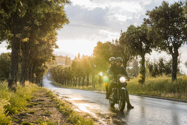 Paar fährt Motorrad auf nasser Straße inmitten von Bäumen bei Sonnenuntergang - CAVF53792