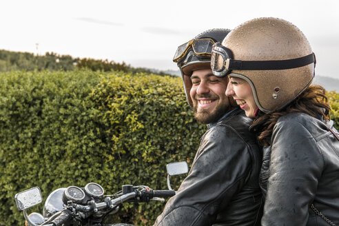 Fröhliches Paar im Gespräch auf dem Motorrad sitzend gegen den Himmel - CAVF53794