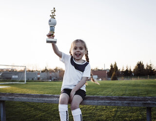 Portrait of happy girl holding trophy while sitting on bench against clear sky at park - CAVF53861