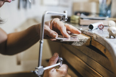 Abgeschnittene Hände einer Kunsthandwerkerin, die eine Handsäge benutzt, während sie auf einem Holztisch in einer Werkstatt Schmuck herstellt - CAVF53904