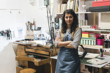 Porträt einer selbstbewussten Kunsthandwerkerin mit verschränkten Armen, die in einer Werkstatt vor einem Regal steht - CAVF53914