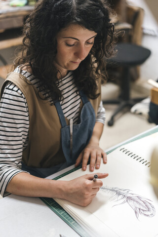 Blick von oben auf eine Kunsthandwerkerin, die in der Werkstatt sitzend an einem Buch zeichnet, lizenzfreies Stockfoto