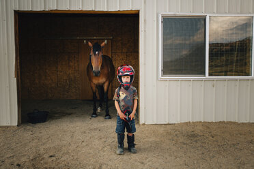 Porträt eines niedlichen Jungen, der einen Helm trägt, während er vor einem Pferd im Stall steht - CAVF53947