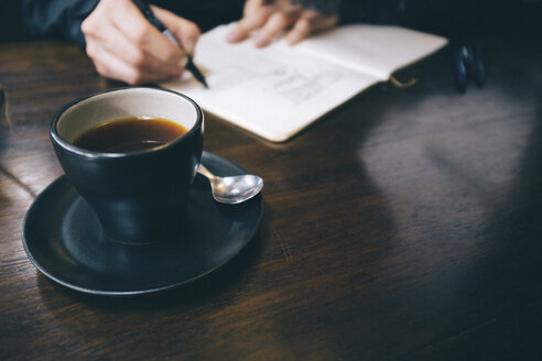 Abgeschnittene Hände eines schreibenden Mannes mit schwarzem Kaffee auf dem Tisch - CAVF53966