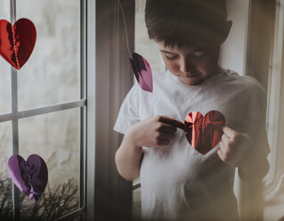 Boy playing with heart shape papers while standing by window at home - CAVF53969