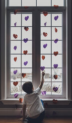 Rear view of boy playing with heart shape decorations by window at home - CAVF53970