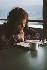 Junge Frau liest Buch mit Kaffeetasse am Tisch - CAVF53977
