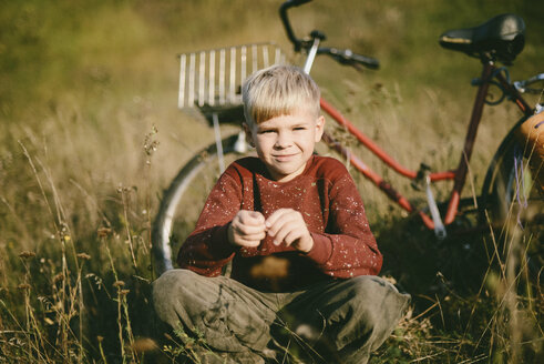 Porträt eines Jungen, der auf einem Feld vor einem Fahrrad hockt - CAVF54001