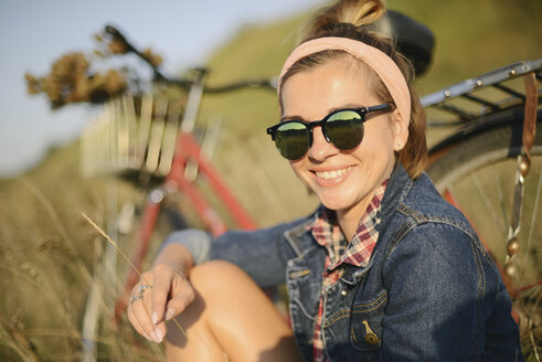 Porträt einer fröhlichen Frau mit Sonnenbrille vor einem Fahrrad auf einem Feld - CAVF54004