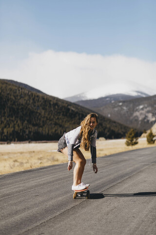 Unbeschwerte junge Frau beim Skateboarden auf der Straße an einem sonnigen Tag in voller Länge, lizenzfreies Stockfoto