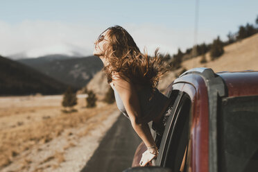 Side view of carefree young woman with tousled hair leaning out from car window - CAVF54033