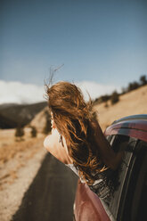 Carefree young woman with tousled hair leaning out from car window - CAVF54034