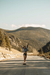 Full length of carefree young woman with arms raised skateboarding on road against mountains - CAVF54035