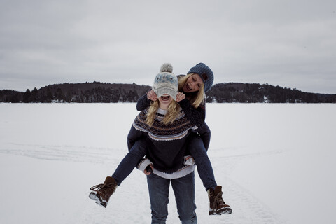 Verspielte Frau, die an der Strickmütze ihrer Freundin zieht, während sie von ihr im Winter im Schnee huckepack genommen wird, lizenzfreies Stockfoto