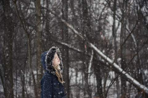 Seitenansicht eines Mädchens mit geschlossenen Augen, das warme Kleidung trägt und bei Schneefall an einem Baum steht, lizenzfreies Stockfoto