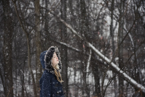 Seitenansicht eines Mädchens mit geschlossenen Augen, das warme Kleidung trägt und bei Schneefall an einem Baum steht - CAVF54086