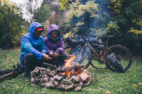 Laughing couple with with bmx bikes sitting at camp fire grilling sausages - OCMF00092