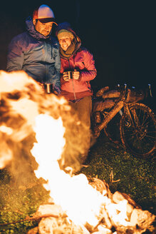 Couple with with bikes standing at camp fire looking at flame by night - OCMF00094