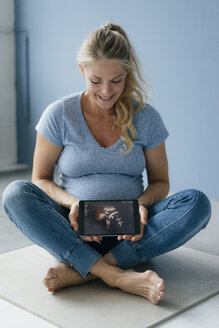 Lächelnde schwangere Frau, die auf dem Boden sitzt und ein Ultraschallbild auf einem Tablet zeigt - KNSF05211