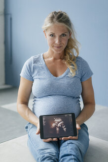Porträt einer lächelnden schwangeren Frau, die auf dem Boden sitzt und ein Ultraschallbild auf einem Tablet zeigt - KNSF05214