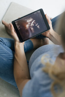 Schwangere Frau sitzt auf dem Boden und betrachtet ein Ultraschallbild auf einem Tablet - KNSF05215