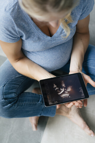 Schwangere Frau sitzt auf dem Boden und betrachtet ein Ultraschallbild auf einem Tablet, lizenzfreies Stockfoto