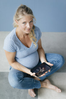 Porträt einer lächelnden schwangeren Frau, die auf dem Boden sitzt und ein Ultraschallbild auf einem Tablet zeigt - KNSF05220