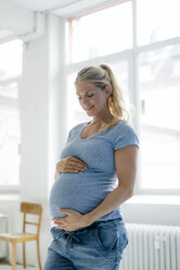 Lächelnde schwangere Frau, die am Fenster steht und auf ihren Bauch schaut - KNSF05234