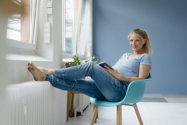 Portrait of smiling pregnant woman sitting on a chair at the window holding tablet - KNSF05249