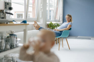 Schwangere Frau sitzt auf einem Stuhl am Fenster mit Kleinkind im Vordergrund - KNSF05252