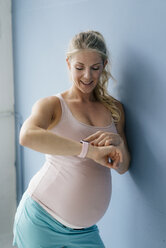 Portrait of smiling pregnant woman standing at blue wall looking at smartwatch - KNSF05260