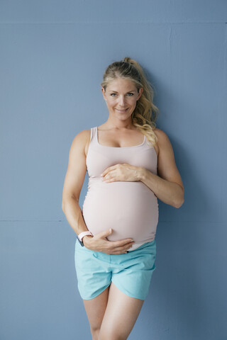 Porträt einer lächelnden schwangeren Frau, die an einer blauen Wand steht, lizenzfreies Stockfoto
