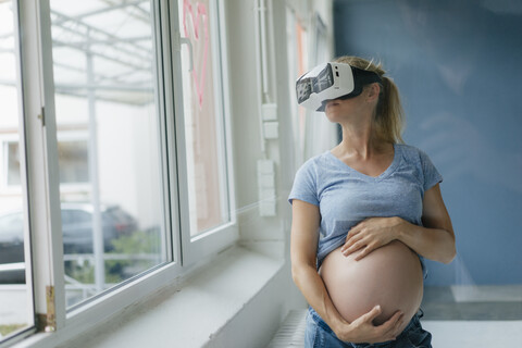 Schwangere Frau mit VR-Brille am Fenster, lizenzfreies Stockfoto