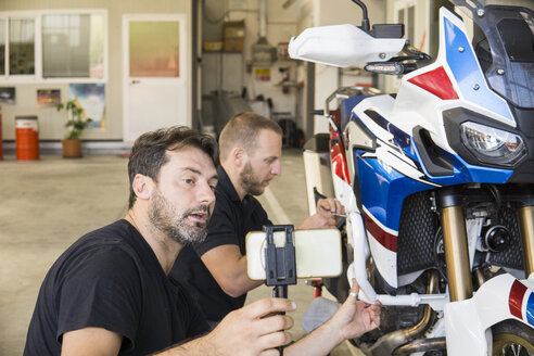 Mechanic working on motorcycle in workshop filmed by his partner - FBAF00154