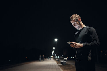 Sportive young man with smartphone and earphones outdoors at night - ZEDF01738