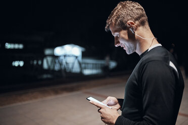 Sportlicher junger Mann mit Smartphone und Kopfhörern im Freien bei Nacht - ZEDF01739