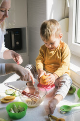 Kleiner Junge sitzt barfuß auf der Arbeitsplatte in der Küche und presst eine Orange aus - MOMF00535