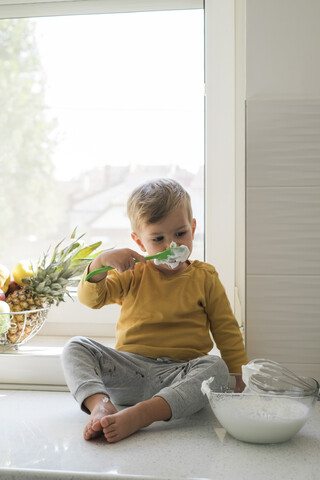 Porträt eines kleinen Jungen, der barfuß auf einer Arbeitsplatte in der Küche sitzt und Schlagsahne nascht, lizenzfreies Stockfoto