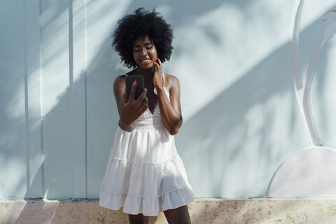 Smiling young woman wearing white dress looking at cell phone at a wall stock photo