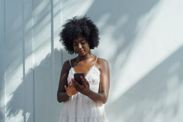 Young woman wearing white dress using cell phone at a wall - BOYF00817
