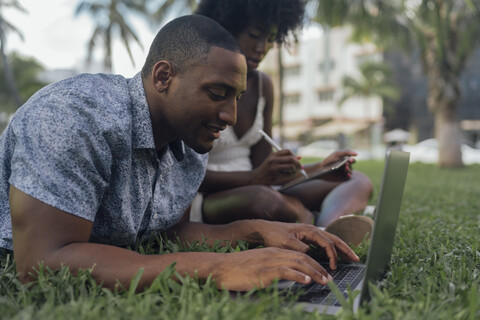 USA, Florida, Miami Beach, junges Paar mit Tablet und Laptop auf Rasen in einem Park, lizenzfreies Stockfoto