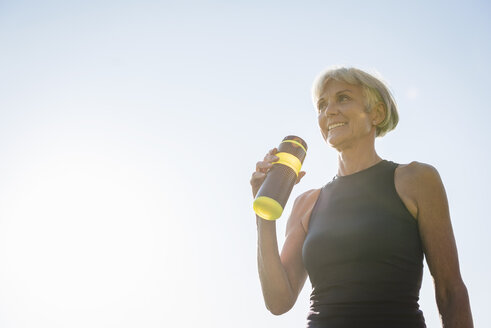 Lächelnde sportliche ältere Frau mit Flasche unter blauem Himmel - DIGF05428