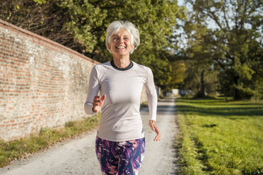 Happy senior woman running along brick wall in a park - DIGF05436