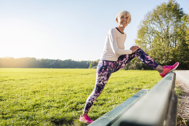 Smiling senior woman stretching on a bench in rural landscape - DIGF05438