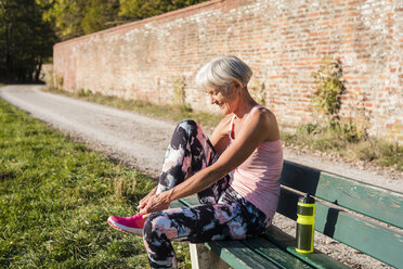 Smiling sportive senior woman sitting on a bench tying her shoes - DIGF05443